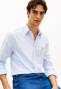 Ανοιχτό μπλε και λευκό πουκάμισο με κουμπιά σε καρό σχέδιο, μακριά μανίκια και μικρό λογότυπο. Φορεμένο με μπλε τζιν παντελόνι. Λεία υφή υφάσματος.