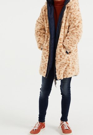 Manteau en fausse fourrure imprimé léopard beige avec doublure marine, fermeture éclair et poches latérales, porté sur un pull à col roulé rouge et un jean bleu foncé.