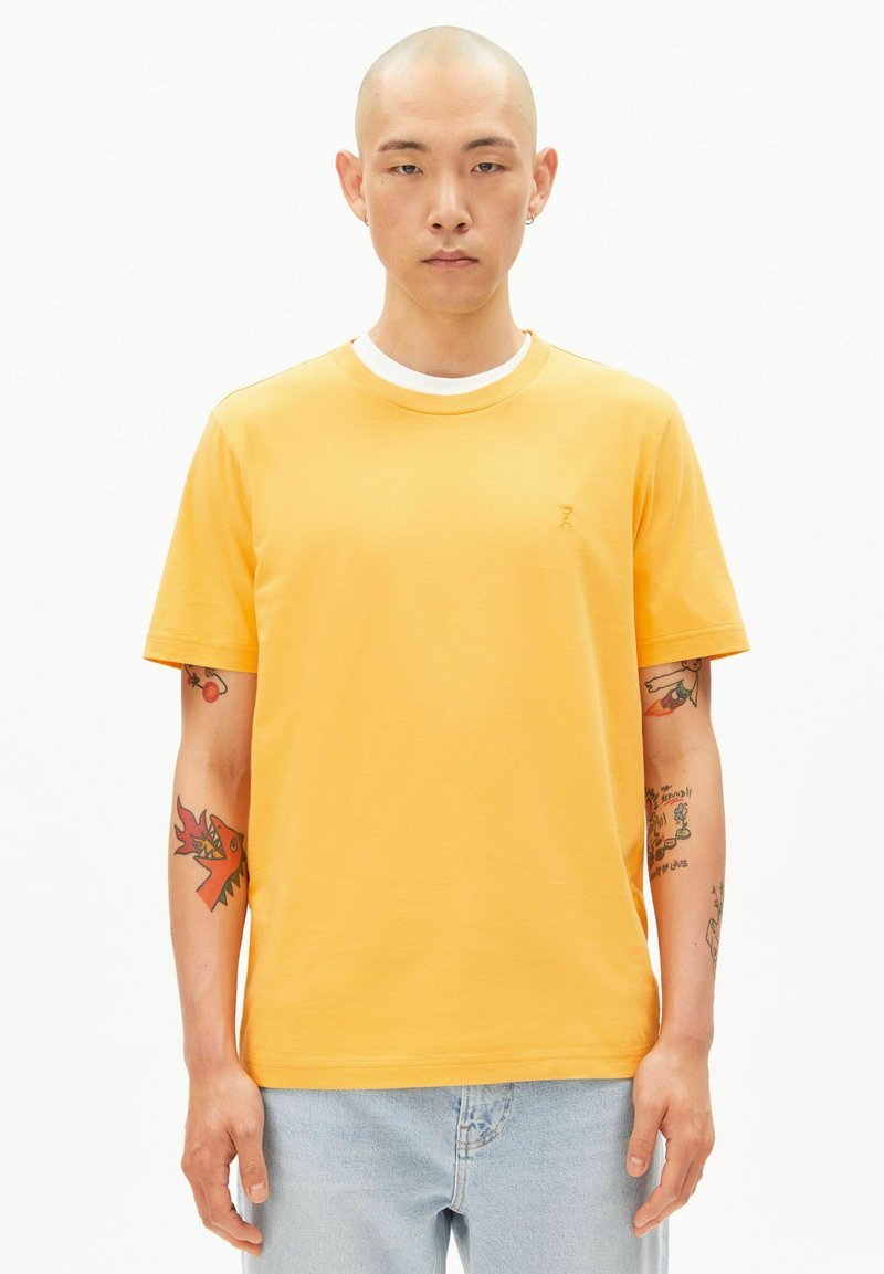 Junger kahlköpfiger Mann, der ein gelbes T-Shirt und hellblaue Jeans trägt, mit sichtbaren Tätowierungen auf beiden Unterarmen, stehend vor einem schlichten weißen Hintergrund.