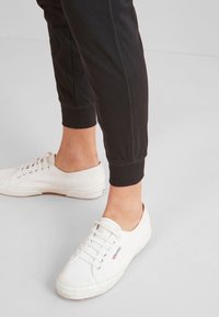 Musta kapeat jogger-housut elastisilla hihnoilla yhdistettynä valkoisiin kangastossuihin, joissa on teksturoitu pinta ja klassinen nauhakiinnitys.