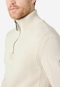 Cremfarbener Strickpullover mit hohem Rippkragen, einem halben Reißverschluss vorne und strukturierten Ärmeln mit geflochtenem Muster. Dezentes Branding-Detail.