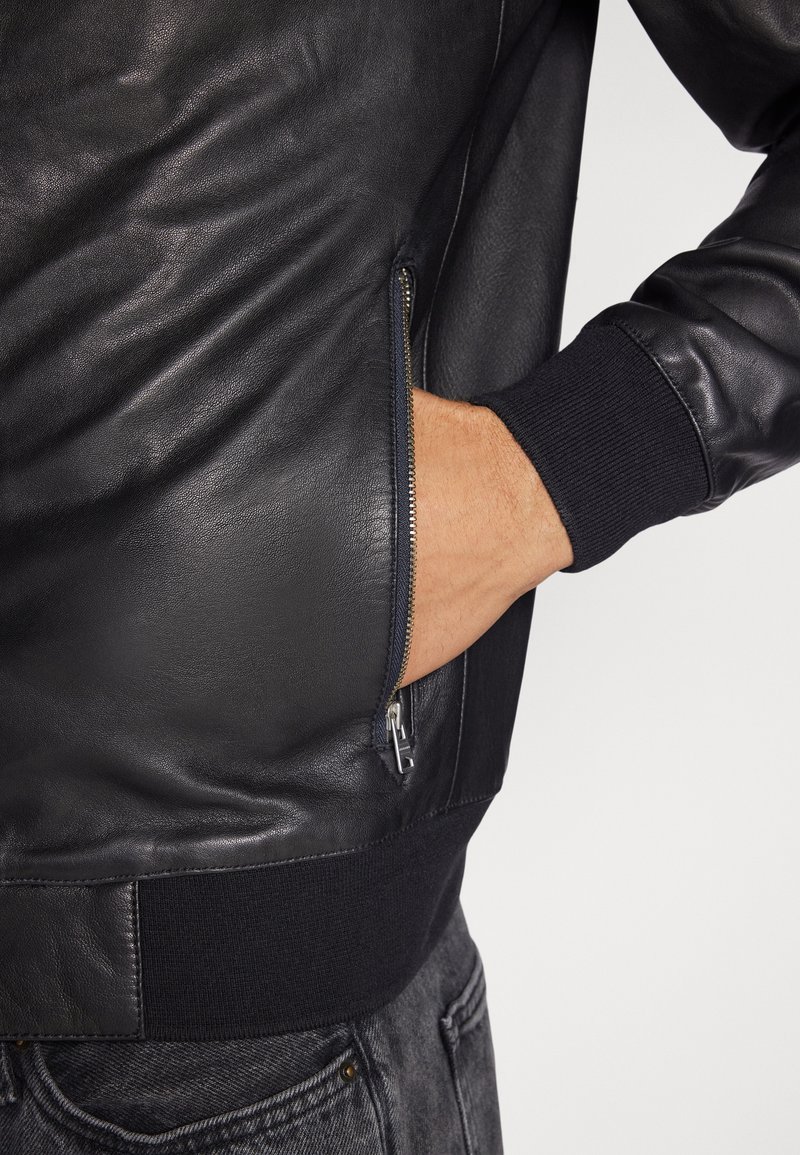 Veste bomber en cuir noir avec des poignets et un ourlet côtelés, dotée d'une poche latérale zippée et d'une finition lisse et texturée.