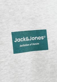 Žalias stačiakampio formos logotipas su baltais užrašais „Jack&Jones“ viršuje ir „Definition of Denim“ apačioje ant šviesiai pilko audinio fono.