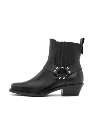 Stivale alla caviglia in pelle nera con tacco basso, pannelli laterali elasticizzati e cinturino decorativo con anello in metallo e borchie su sfondo bianco.