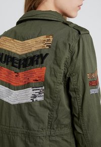 Chaqueta de algodón verde con franjas de zigzag bordadas en oro, naranja, blanco y negro. Incluye el texto "SUPERDRY" en la parte trasera.