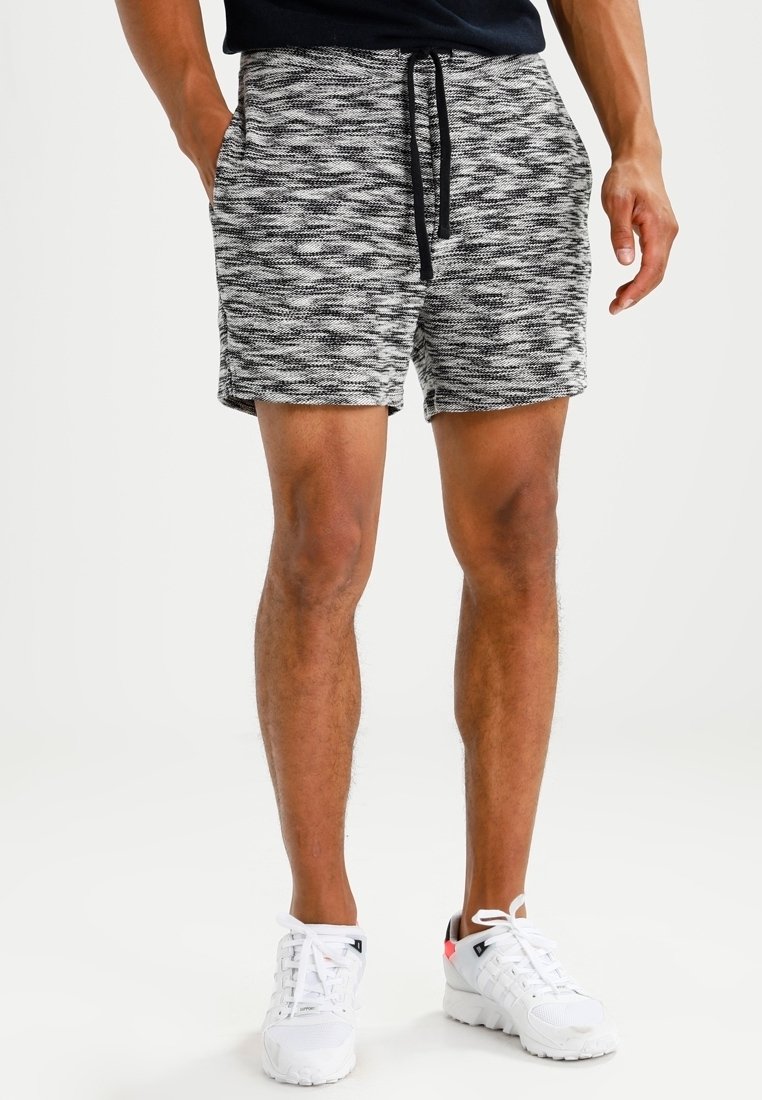 Homme portant un short tricoté noir et blanc avec un cordon de serrage noir, une chemise noire et des baskets blanches, les mains dans les poches.