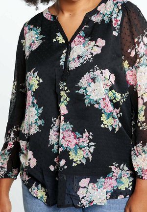 Vrouw die een zwarte doorschijnende blouse draagt met roze, witte, gele en groene bloemenpatronen en driekwart mouwen.