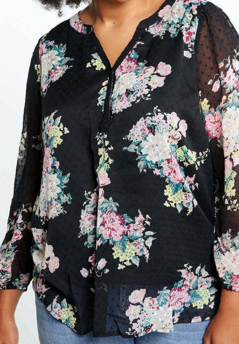 Femme portant un chemisier noir transparent avec des motifs floraux roses, blancs, jaunes et verts et des manches trois-quarts.