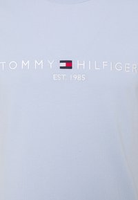 Tela azul claro con texto bordado en blanco "TOMMY HILFIGER" y el logo de la marca con la bandera roja, blanca y azul encima "EST. 1985".