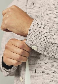 Textured light gray jacket with a woven pattern, featuring a buttoned cuff, and a white button accent. Close-up of hand adjusting sleeve.