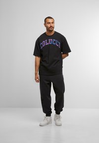 Černé oversized tričko s nápisem "COLUCCI" v barevných písmenech, spárované s černými tepláky a bílými teniskami. Hladká tkanina, volný střih.