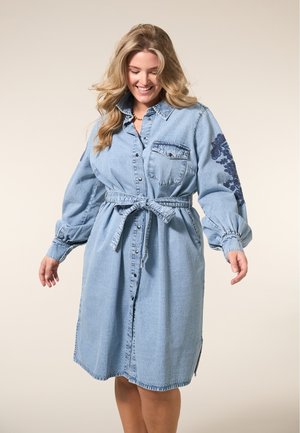 Lichtblauwe denim shirtjurk met lange pofmouwen, een ceintuur in de taille en een voorzak. Kenmerkt zich door donkere bloemenborduursels op één mouw.