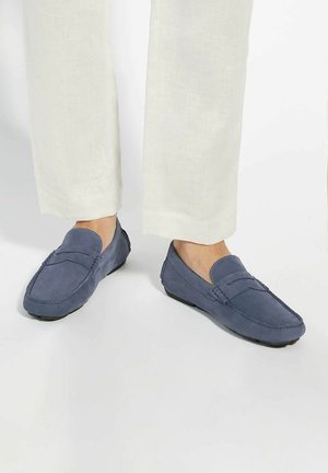 Blue suede loafers with a flat sole, featuring a stitched design and a decorative band across the upper. Worn with light-colored pants.