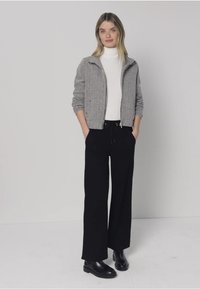 Harmaa vetoketjullinen takki, jossa on teksturoitu kangas, valkoinen pooloneule ja mustat leveälahkeiset housut. Malli seisoo käsineet taskuissa ja pukeutuneena mustiin kenkiin.