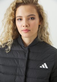 Crna puffer jakna s visokim ovratnikom i horizontalnim šavovima. Istaknuti bijeli Adidas logo na lijevoj strani prsnog područja.