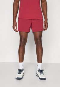 Red athletic shorts with a smooth texture, featuring a simple design and a folded hem. Paired with white socks and navy and white sneakers.