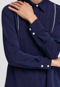 Blusa blu navy con maniche lunghe, chiusura anteriore con bottoni e dettagli in bianco. Presenta bottoni rotondi di tipo perla sui polsini. Tessuto morbido.