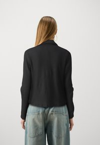 Černý texturovaný blazer s dlouhými rukávy, se zářezovým límcem, spárovaný se světle modrými džínami s vysokým pasem. Zadní pohled.