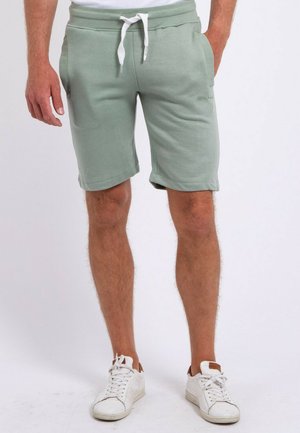 Homme portant un short vert clair à cordon et des baskets blanches, debout devant un fond uni.