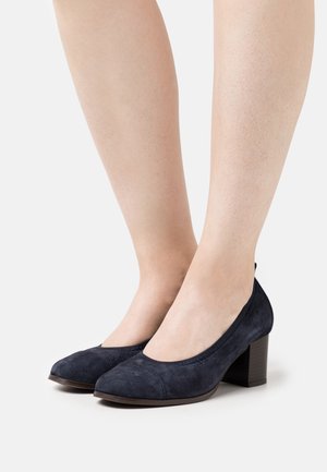 Scarpe col tacco alto in suede blu navy indossate da gambe nude, con una ripresa ravvicinata delle calzature e delle caviglie su uno sfondo bianco semplice.