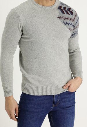 Mężczyzna w jasnoszarym swetrze dzianinowym z wielokolorowym geometrycznym wzorem na lewym ramieniu, w połączeniu z ciemnoniebieskimi dżinsami.