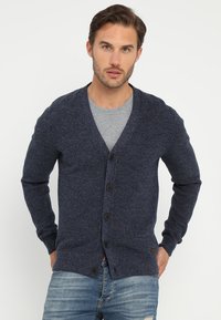 Marinblå stickad cardigan med V-ringning, fem knappar och långa ärmar. Noterbart strukturerat mönster och en liten logotypdetalj vid fållen.
