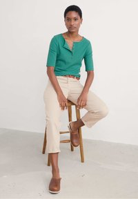 Zeleni rebrasti top s kratkim rukavima i dugmadi, uparen s bež uskim hlačama i smeškim kožnim klocama, sjedi na drvenom tabureu.