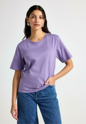 Kvinde med langt mørkt hår iført sølvfarvede øreringe i hoop-stil, en lavendel kortærmet bluse og blå jeans, poserer foran en ensfarvet lys baggrund.