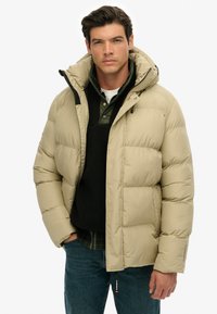 Beige puffjakke med kviltet design, der har hætte, sorte detaljer og lynlåslukning foran. Laget over en mørk pullover og en ternet skjorte.