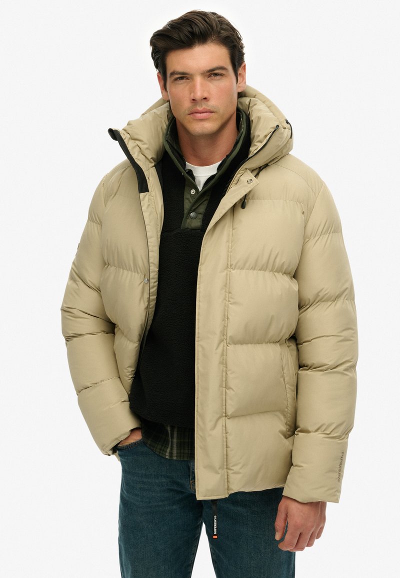 Beige puffjakke med kviltet design, der har hætte, sorte detaljer og lynlåslukning foran. Laget over en mørk pullover og en ternet skjorte.