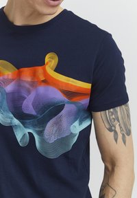 Tummansininen puuvillapaita, jossa on värikäs graafinen kuvio, jossa on aaltoilevia viivoja oranssina, violetina ja akvana. Lyhyet hihat ja pyöreä kaula-aukko.