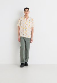 Camisa de manga corta en crema con estampado de frutas, combinada con pantalones holgados en un verde apagado y zapatos negros, de pie frente a un fondo blanco.
