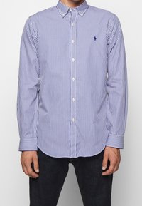 Mies, der ein langärmliges, blau-weiß gestreiftes Button-Up-Hemd mit einem kleinen gestickten Logo auf der linken Brust trägt.