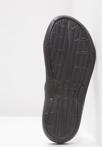 Semelle de chaussure en caoutchouc noir avec un motif grip ondulé texturé et le nom de marque "Crocs" embossé au centre.