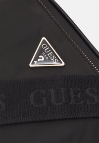 Czarna nylonowa torba z trójkątnym metalowym logo z napisem "GUESS U.S.A." oraz znakiem zapytania, a także teksturowanym nylonowym paskiem z wytłoczonym napisem "GUESS."