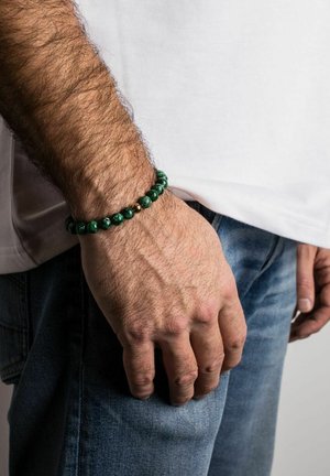 Zöld gyöngy karkötő, texturált felülettel és arany részletekkel, viselve egy csuklón, fehér inggel és kék farmernadrággal párosítva.