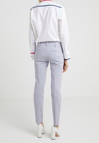 Světle šedé kalhoty na míru s úzkým střihem, mají zadní kapsy a boční švy. Oblečeno s bílou košilí, zapnutým límečkem a červenými akcenty.