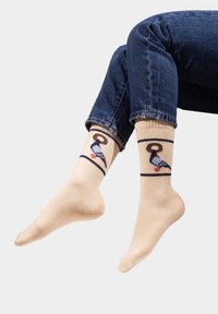 Beżowe skarpetki z prążkowaną teksturą, zdobione wzorem kolorowych ptaków. Projekt zawiera ciemny kontur i czerwone akcenty na stopach ptaków.