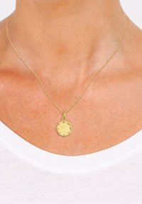Collier chaîne en or avec un pendentif rond texturé porté autour du cou d'une personne sur un fond de chemise blanche.