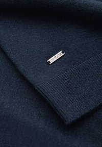 Dunkelblau gestrickter Pullover mit gerippten Bündchen, ausgestattet mit einem silbernen Metallschild mit der Aufschrift "ombre", das am Saum angebracht ist.