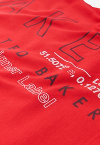 Camiseta de algodón roja con texto impreso en negrita en blanco y negro; los detalles incluyen coordenadas geográficas y el nombre de la marca.