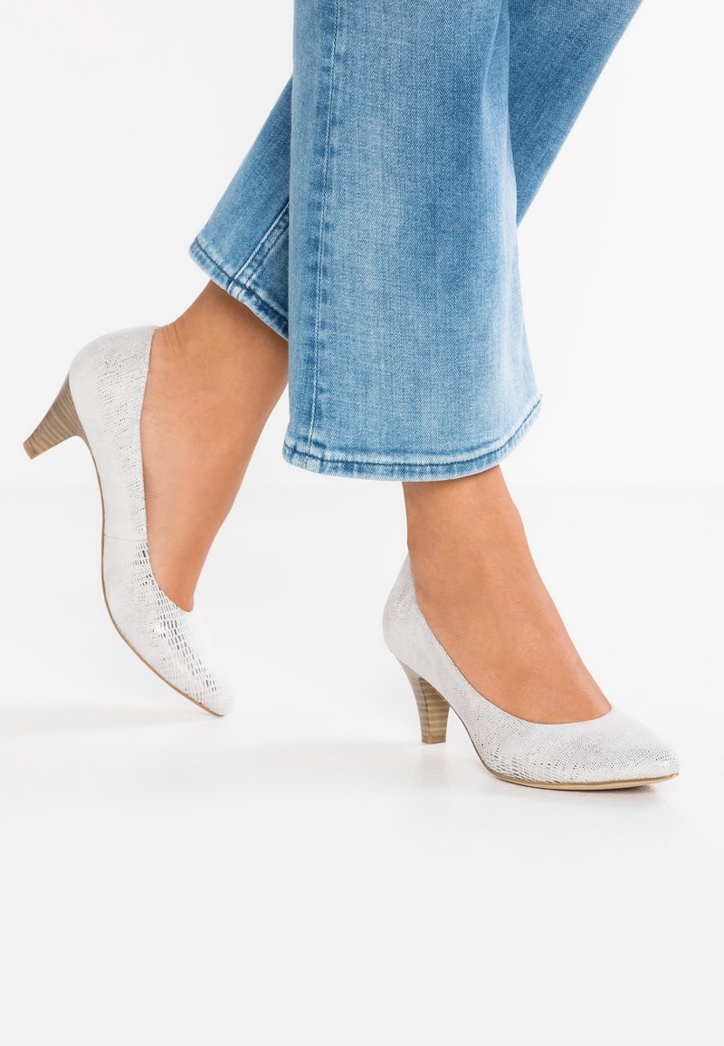 Pieds portant un jean bleu clair et des chaussures blanches texturées à petit talon, un pied légèrement levé sur fond blanc.