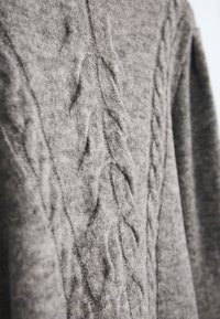 Gros pull en tricot gris avec un motif en tresses. Texture douce, manches longues et poignets côtelés. Le tissu semble chaud et épais.