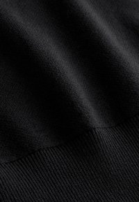 Közeli kép fekete, texturált kötött anyagról, bordázott szegéllyel, finom szövést és puha anyagredőket mutatva.