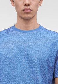 Νεαρός άνδρας φορά μπλε T-shirt με στρογγυλή λαιμόκοψη και επαναλαμβανόμενο ανοιχτό μπλε μοτίβο "MK" στο ύφασμα, εμφανιζόμενος από τους ώμους μέχρι τα χείλη.