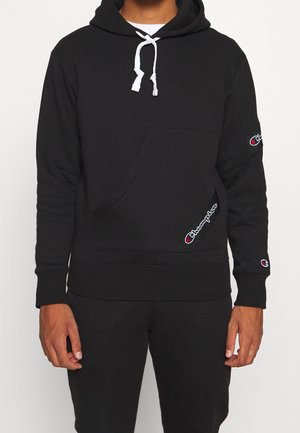 Mann trägt schwarzen Champion-Hoodie mit weißen Kordeln und Logo-Aufnähern an Tasche und Ärmeln, kombiniert mit passenden schwarzen Hosen.