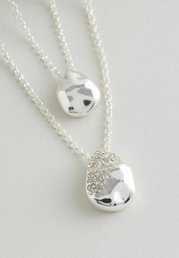 Sudraba toņa divkāršā kaklarota ar diviem teksturētiem, pulētiem piekariņiem; viens rotāts ar caurspīdīgiem rhinestones uz smalkas ķēdītes.