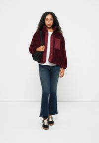 Bordeaux fleece takki, jossa on neppari etuosassa, rintatasku ja pehmeä rakenne, yhdistettynä sinisiin leveälahkeisiin farkkuihin ja mustiin lenkkareihin.