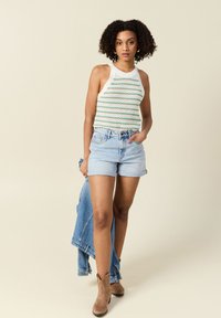 Mouwloze gebreide top met groene en witte strepen, gecombineerd met lichte denimshorts. Bruine enkellaarzen maken de outfit af, met een denimjack in de hand.