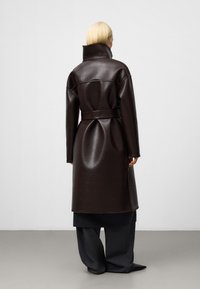 Cappotto in faux leather marrone con colletto alto, vita cintata e vestibilità oversize, caratterizzato da una texture liscia e maniche lunghe.
